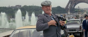 Movie still from “The Brain” (1969), directed by Gérard Oury – An older man in a gray jacket and hat holding a camera; Medium shot, Low angle