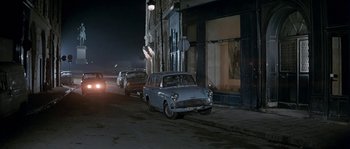 Movie still from “The Brain” (1969), directed by Gérard Oury – An old car parked on the side of the street; Wide shot, High angle
