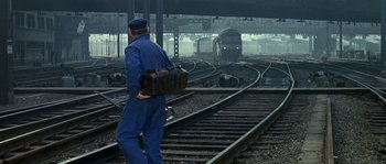 Movie still from “The Brain” (1969), directed by Gérard Oury – A man in a blue coverall is standing on train tracks holding a bag; Wide shot, High angle