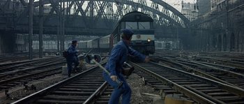 Movie still from “The Brain” (1969), directed by Gérard Oury – A man in blue overalls is walking on train tracks while another man in blue overalls is holding a wrench; Wide shot, High angle