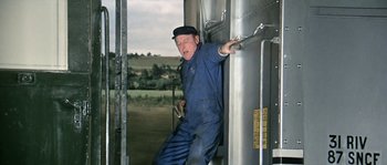 Movie still from “The Brain” (1969), directed by Gérard Oury – A man in blue coveralls leaning against a metal tank; Medium shot, Low angle
