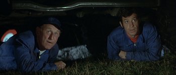 Movie still from “The Brain” (1969), directed by Gérard Oury – A couple of men laying in the grass under a car; Medium shot, Low angle