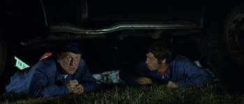 Movie still from “The Brain” (1969), directed by Gérard Oury – Two men laying in the grass under a car; Medium shot, Low angle