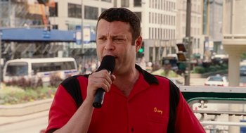 Movie still from “The Break-Up” (2006), directed by Peyton Reed – A man in a red shirt is holding a microphone; Close Up shot, Low angle