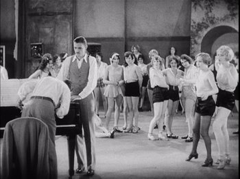 Movie still from “The Broadway Melody” (1929), directed by Harry Beaumont – A group of people standing in a room with a piano; Wide shot, High angle