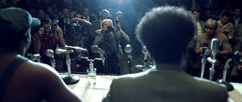 Movie still from “Ali” (2001), directed by Michael Mann – A man taking a picture of a crowd of people in front of microphones and microphones; Extreme Close Up shot, Over the shoulder angle
