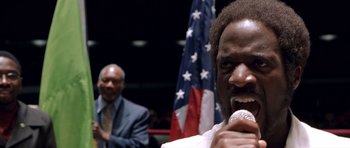 Movie still from “Ali” (2001), directed by Michael Mann – A man with a microphone in front of an american flag; Close Up shot, Low angle