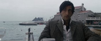 Movie still from “The Brothers Bloom” (2008), directed by Rian Johnson – A man sitting at a table holding a coffee mug; Close Up shot, Over the shoulder angle
