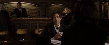 Movie still from “The Brothers Bloom” (2008), directed by Rian Johnson – A man sitting at a table in a suit and tie; Medium shot, Over the shoulder angle