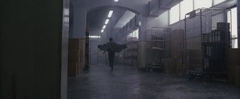 Movie still from “The Brothers Bloom” (2008), directed by Rian Johnson – A man walking in a warehouse holding a surfboard in the rain; Extreme Wide shot, High angle