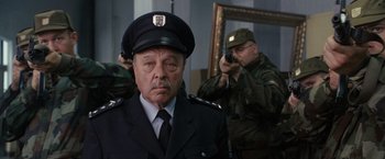 Movie still from “The Brothers Bloom” (2008), directed by Rian Johnson – A man in a police uniform with a gun in front of him; Medium shot, Over the shoulder angle