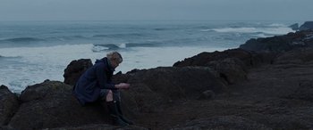 Movie still from “The Burning Plain” (2008), directed by Guillermo Arriaga – A woman sitting on top of a rock near the ocean; Wide shot, High angle
