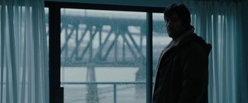 Movie still from “The Burning Plain” (2008), directed by Guillermo Arriaga – A man standing in front of a window with a bridge in the background; Medium shot, Over the shoulder angle