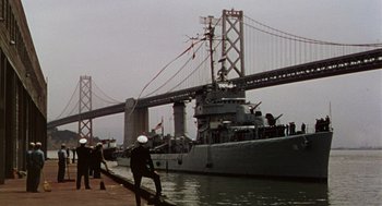 Movie still from “The Caine Mutiny” (1954), directed by Edward Dmytryk – A man standing next to a boat on a body of water near a bridge; Extreme Wide shot, Low angle