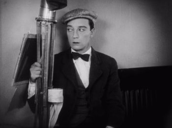 Movie still from “The Cameraman” (1928), directed by Buster Keaton – A man in a suit and hat holding an umbrella in front of a wall; Medium shot, Low angle