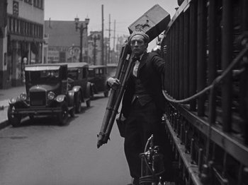Movie still from “The Cameraman” (1928), directed by Buster Keaton – A man standing next to a fence holding a bunch of suitcases; Wide shot, Low angle