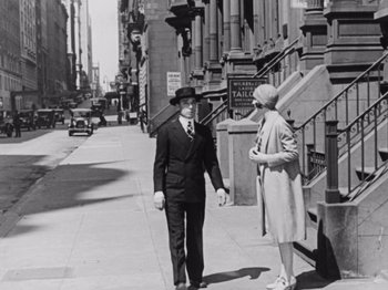 Movie still from “The Cameraman” (1928), directed by Buster Keaton – A man in a suit talking to a woman on the sidewalk; Wide shot, Low angle