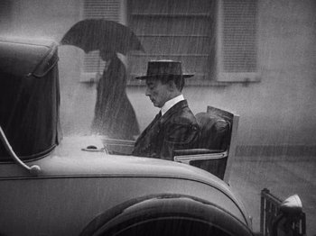 Movie still from “The Cameraman” (1928), directed by Buster Keaton – A man sitting in the back of an antique car in the pouring rain with an umbrella; Wide shot, Over the shoulder angle