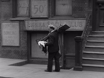 Movie still from “The Cameraman” (1928), directed by Buster Keaton – A black and white photo of a man carrying a bunch of suitcases; Wide shot, High angle