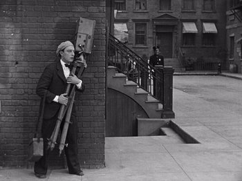 Movie still from “The Cameraman” (1928), directed by Buster Keaton – An old man leaning against a brick wall holding a camera; Wide shot, Low angle
