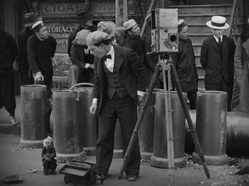 Movie still from “The Cameraman” (1928), directed by Buster Keaton – A man in a black suit and a hat is standing next to an old camera; Wide shot, High angle
