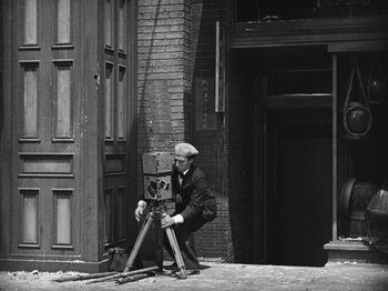 Movie still from “The Cameraman” (1928), directed by Buster Keaton – An old man is standing next to an old camera; Wide shot, High angle