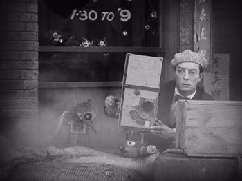 Movie still from “The Cameraman” (1928), directed by Buster Keaton – A man holding a camera in front of a window; Medium shot, High angle