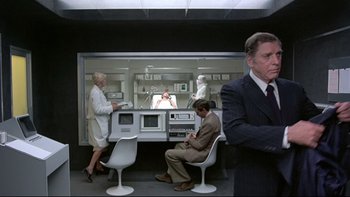 Movie still from “The Cassandra Crossing” (1976), directed by George P. Cosmatos – A group of people sitting at a table in front of a microwave oven; Wide shot, Over the shoulder angle
