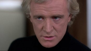 Movie still from “The Cassandra Crossing” (1976), directed by George P. Cosmatos – An older man with blonde hair wearing a black shirt; Close Up shot, Over the shoulder angle