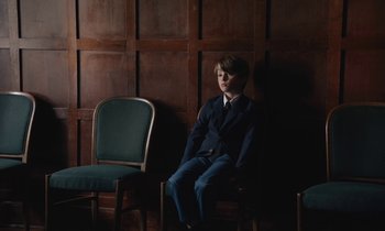 Movie still from “The Cathedral” (2021), directed by Ricky D'Ambrose – A boy in a suit sitting in a chair; Medium shot, High angle