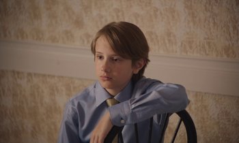 Movie still from “The Cathedral” (2021), directed by Ricky D'Ambrose – A young man sitting in a chair wearing a shirt and tie; Close Up shot, High angle