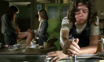 Movie still from “The Cement Garden” (1993), directed by Andrew Birkin – A woman sitting at a kitchen sink while another woman sits on a stool; Medium shot, Low angle