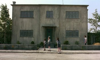 Movie still from “The Cement Garden” (1993), directed by Andrew Birkin – A group of people standing in front of a building on the sidewalk; Extreme Wide shot, Low angle