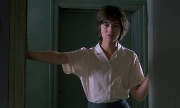 Movie still from “The Cement Garden” (1993), directed by Andrew Birkin – A woman in white shirt and black skirt standing in front of a door; Medium shot, Low angle