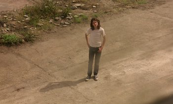 Movie still from “The Cement Garden” (1993), directed by Andrew Birkin – A person standing on a dirt road with their hands on their hips; Wide shot, High angle