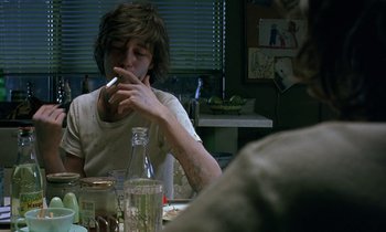Movie still from “The Cement Garden” (1993), directed by Andrew Birkin – A man sitting at a table smoking a cigarette; Medium shot, Over the shoulder angle
