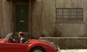 Movie still from “The Cement Garden” (1993), directed by Andrew Birkin – A man driving a red car down a street; Wide shot, Over the shoulder angle