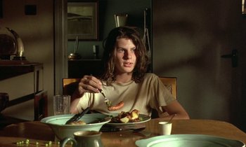Movie still from “The Cement Garden” (1993), directed by Andrew Birkin – A person sitting at a table eating food; Medium shot, Low angle