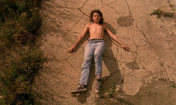 Movie still from “The Cement Garden” (1993), directed by Andrew Birkin – A man laying on the ground with no shirt on; Wide shot, Overhead angle
