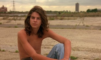 Movie still from “The Cement Garden” (1993), directed by Andrew Birkin – A young man sitting on the ground with his hands on his knees; Close Up shot, Low angle