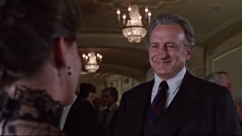 Movie still from “The Changeling” (1980), directed by Peter Medak – An older man wearing a suit and tie smiling; Medium shot, Over the shoulder angle