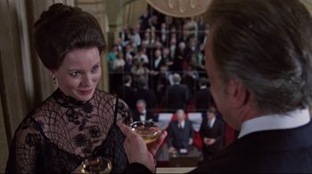 Movie still from “The Changeling” (1980), directed by Peter Medak – A man and a woman are holding wine glasses in front of an audience; Medium shot, Over the shoulder angle