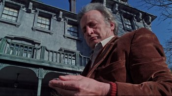 Movie still from “The Changeling” (1980), directed by Peter Medak – An older man in a corduroy jacket holding a cell phone in front of a building; Medium shot, Low angle