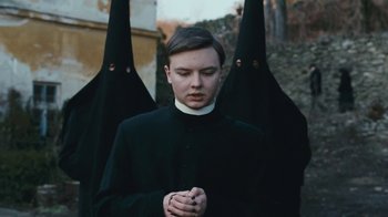 Movie still from “The Childhood of a Leader” (2015), directed by Brady Corbet – A man in a priest's outfit with two black hoods behind him; Medium shot, Low angle