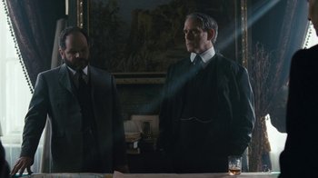 Movie still from “The Childhood of a Leader” (2015), directed by Brady Corbet – Two men standing in front of a painting in a dark room; Medium shot, Over the shoulder angle