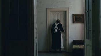 Movie still from “The Childhood of a Leader” (2015), directed by Brady Corbet – A woman in a black dress and white apron standing in front of an open door; Wide shot, High angle