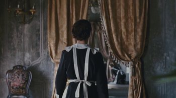 Movie still from “The Childhood of a Leader” (2015), directed by Brady Corbet – A woman in a black and white dress standing in front of a window; Medium shot, Over the shoulder angle