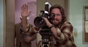 Movie still from “The China Syndrome” (1979), directed by James Bridges – A man holding a camera up to the camera while standing in front of a tripod; Close Up shot, Low angle