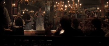 Movie still from “The Claim” (2000), directed by Michael Winterbottom – People are sitting at tables in front of chandeliers; Wide shot, Over the shoulder angle