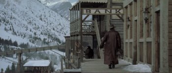 Movie still from “The Claim” (2000), directed by Michael Winterbottom – A man in a long brown coat and hat walking up to a bank building; Wide shot, Low angle